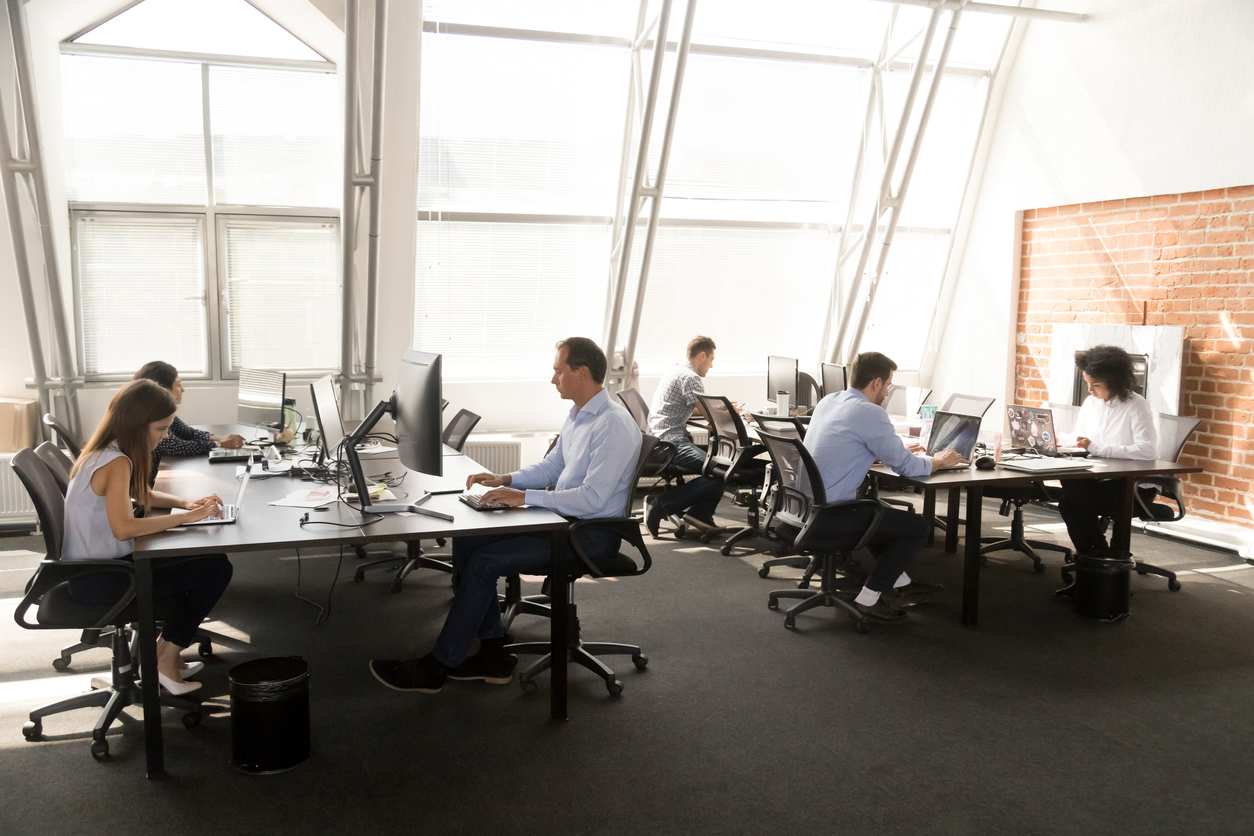 Diverse multiracial workers sitting at desk working in coworking space. Busy six businesspeople coworkers using computers typing chatting solving business matters in modern light contemporary office