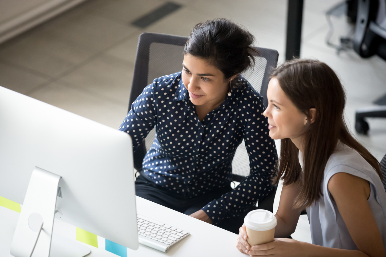 Multiracial colleagues indian and caucasian young women having coffee break sitting together at desk in office. Diverse students interns friends looks at pc screen talking discussing working moments