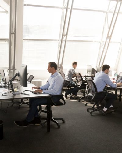 Diverse multiracial workers sitting at desk working in coworking space. Busy six businesspeople coworkers using computers typing chatting solving business matters in modern light contemporary office