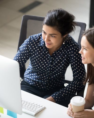 Multiracial colleagues indian and caucasian young women having coffee break sitting together at desk in office. Diverse students interns friends looks at pc screen talking discussing working moments